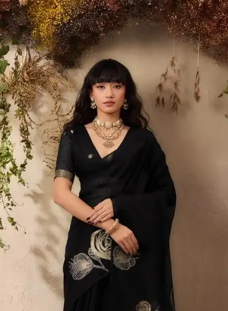 Woman in a black saree with floral patterns standing against a decorative background with plants.