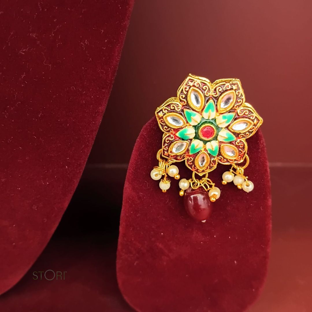 Decorative gold earring with green and red accents on a red velvet stand.
