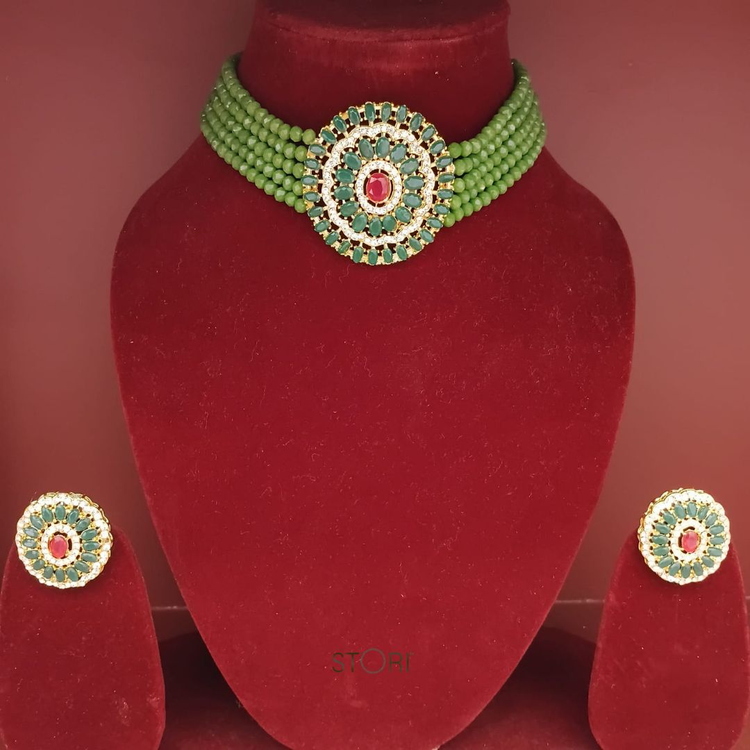 Green beaded necklace with red gemstone on a red velvet stand, accompanied by matching earrings.