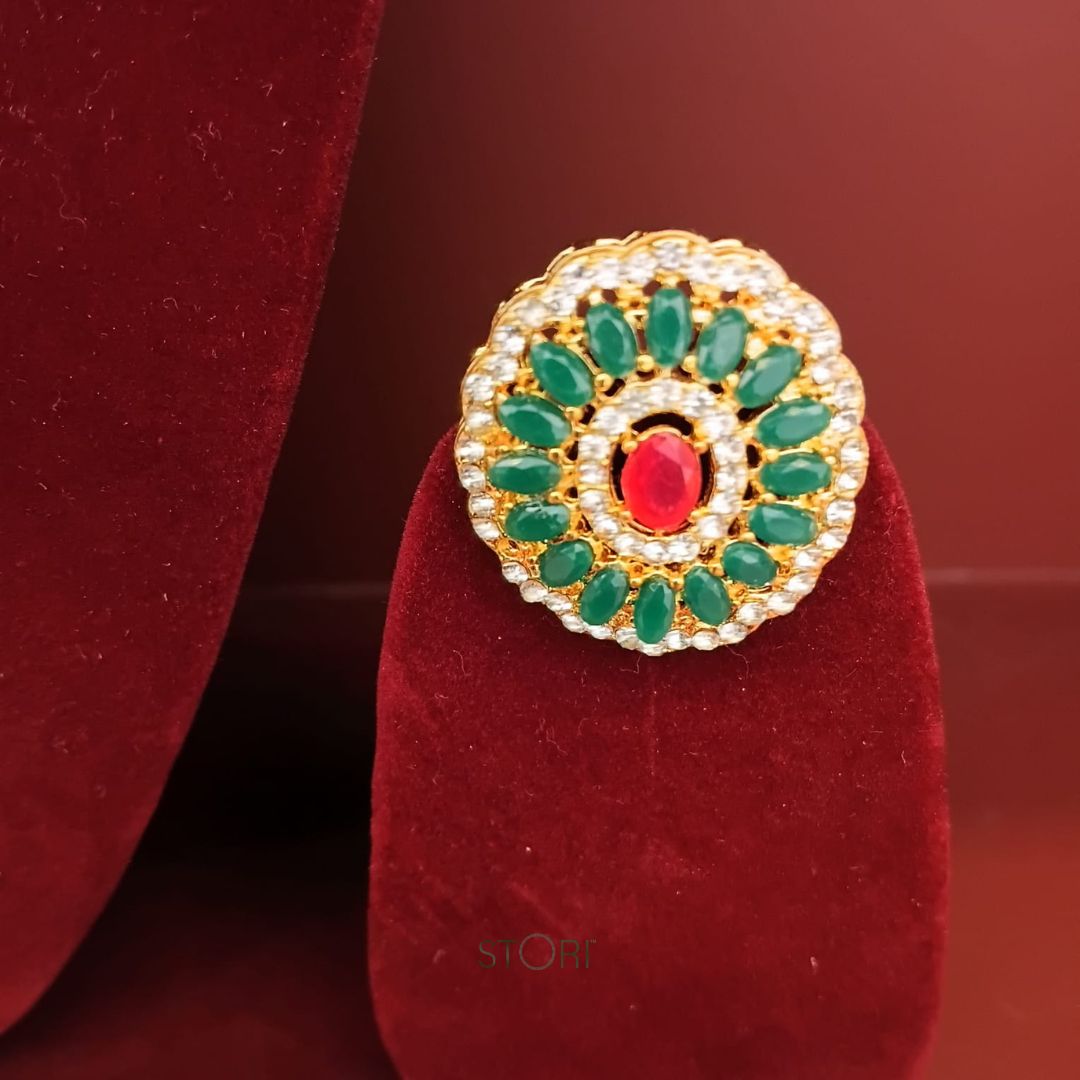 Gold ring with green gemstones and a red stone on a red velvet stand.