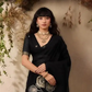 Woman in a black saree with floral patterns standing against a decorative background with plants.