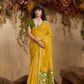 Woman in a yellow saree with green blouse standing against a natural backdrop.