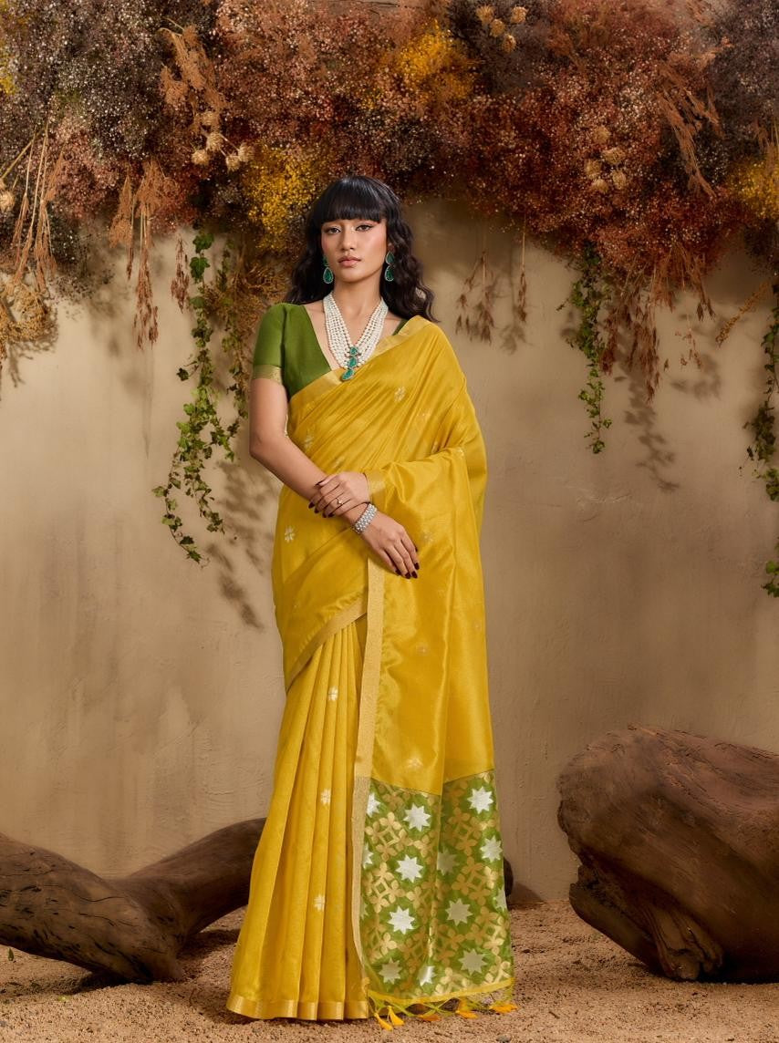 Woman in a yellow saree with green blouse standing against a natural backdrop.