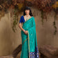 Woman in a teal saree with blue border standing against a natural background