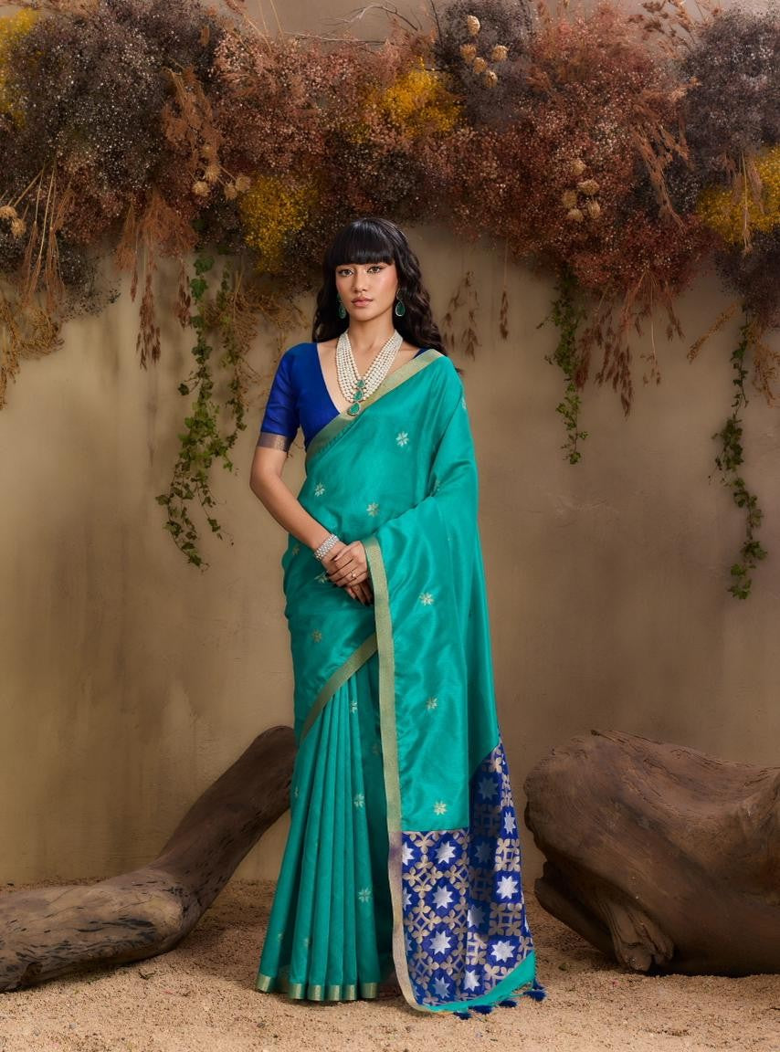 Woman in a teal saree with blue border standing against a natural background