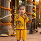A toddler stands barefoot in a decorated temple courtyard, holding a flute. The child is wearing a bright yellow traditional outfit with a printed graphic on the front and brown patterned trim.